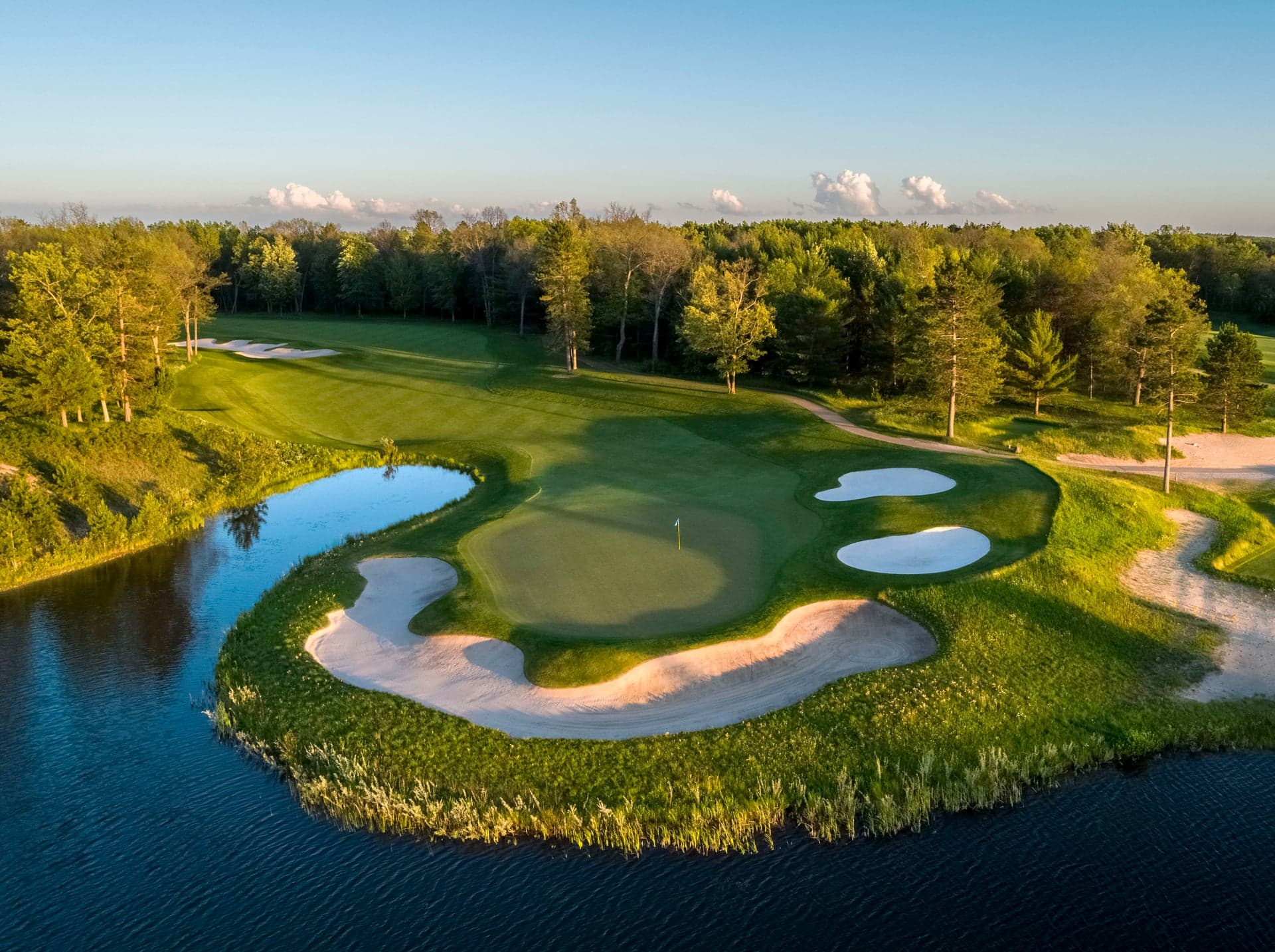 Aerial view of a Forest Dunes green in Roscommon, Michigan, ringed by sand bunkers on a peninsula above a pond fringed with tall native grasses, pine forest beyond in late afternoon light.