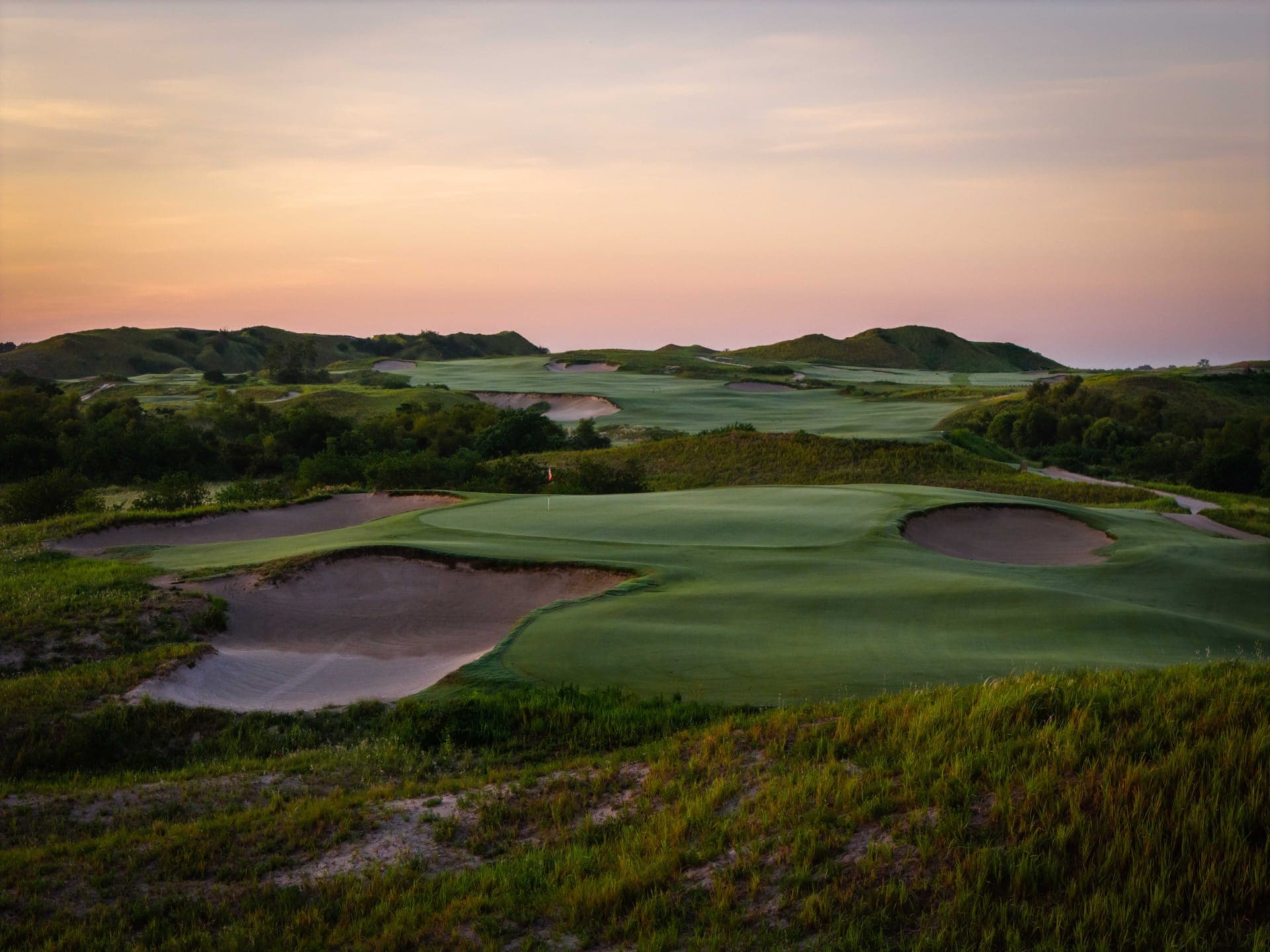 Streamsong Red green at dusk with deep pot bunkers and rolling sand ridges under a pink sky, designed by Coore and Crenshaw